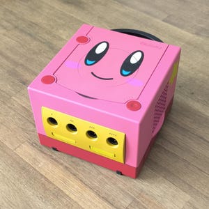 May include: A pink Nintendo GameCube console customized with a Kirby face design. The console features a yellow front panel with controller ports, red accents, and a black handle. The top of the console has a smiling Kirby face.
