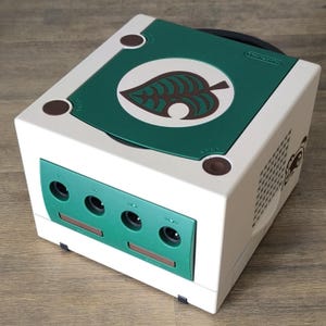 May include: A white and green Nintendo GameCube console with a leaf design on the top. The console has a green front panel with four controller ports and a brown leaf logo. The console is a retro gaming item.