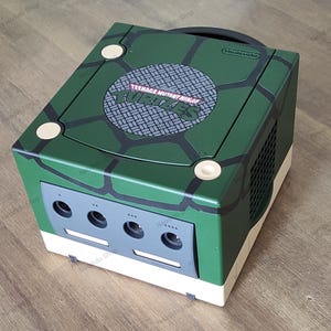 Könnte beinhalten: Eine grüne und weiße Nintendo GameCube Konsole mit einem Teenage Mutant Ninja Turtles Design. Die Konsole hat ein schwarzes Disc-Laufwerk und eine schwarze Lüftung an der Seite. Die Konsole steht auf einer Holzoberfläche.