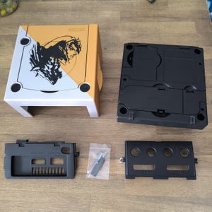 Peut inclure: Une console Nintendo GameCube personnalisée, aux couleurs or et blanc. La console présente un graphisme noir et blanc d'un personnage. Plusieurs composants noirs et un petit outil sont également affichés.