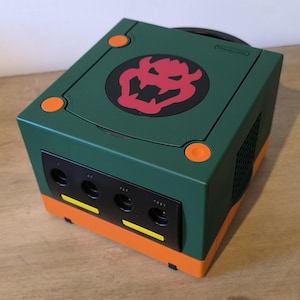 May include: A custom-painted Nintendo GameCube console in green and orange. The top features a Bowser logo in red and black. The console has four controller ports and a disc tray. The power and reset buttons are orange.