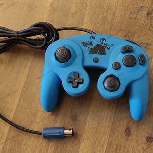 May include: A blue video game controller with a black cable and connector. The controller has a D-pad, analog stick, and buttons. A black graphic of two figures with guns is on the controller. The controller is on a wooden surface.