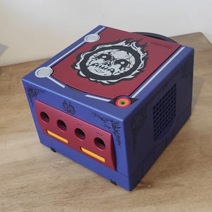 Può includere: Una console Nintendo GameCube personalizzata con uno schema di colori rosso e blu. La parte superiore della console presenta una grafica di teschio in bianco e nero con fiamme. La console ha un design in bianco e nero sui lati.