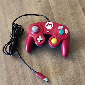 Può includere: Un controller per videogiochi rosso con un cavo nero. Il controller presenta un logo di Mario bianco e pulsanti colorati. Il joystick è grigio e il pad direzionale è una croce bianca.