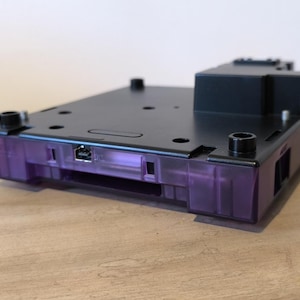 Może przedstawiać: Czarna konsola Nintendo GameCube z przezroczystą, fioletową obudową. Konsola ma gniazdo na kartridż z grą i port na kartę pamięci.