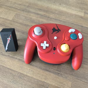 Peut inclure: Manette de jeu vidéo rouge avec des boutons blancs, bleus et jaunes. Une boîte noire avec un graphisme blanc et rouge est à côté de la manette. La manette a un joystick blanc et une croix directionnelle.