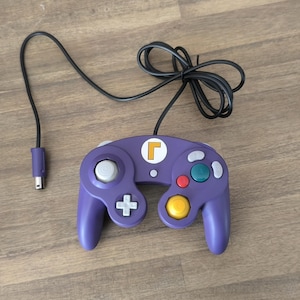 Può includere: Controller GameCube viola con un adesivo bianco con la lettera 'L' sulla parte superiore. Il controller ha un cavo nero.