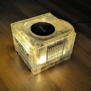 May include: A transparent, illuminated Nintendo GameCube console. The console is lit from within, highlighting its internal parts. The top has a black disc drive cover with the GameCube logo. It rests on a wooden surface.