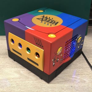 May include: A custom-painted video game console with a vibrant design. The console is a mix of purple, blue, red, and green. The top has a gold emblem with the text "ZELDA". The side features a cartoon graphic of a character.