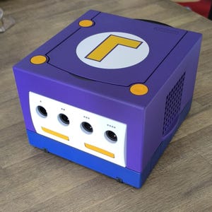 May include: A purple and blue Nintendo GameCube console with a white and yellow L logo on the top. The console has three controller ports and a ventilation grill on the side.