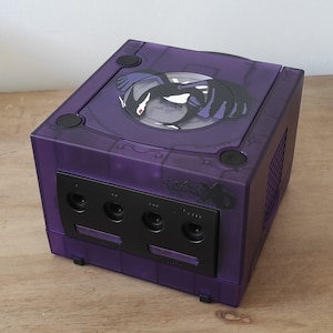 May include: A purple GameCube console with a transparent top and a black and white graphic of a creature. The console has a black front panel with controller ports and the text "Pokemon XD" is visible.