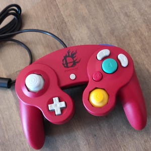 Könnte beinhalten: Ein roter Nintendo GameCube-Controller mit einem weißen C-Stick, einem weißen Steuerkreuz und einer gelben und blauen Tastenanordnung. Der Controller hat ein Super Smash Bros.-Logo mit einem Flammen-Design auf der Oberseite.