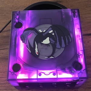Może przedstawiać: Fioletowa konsola GameCube z przezroczystą obudową z tworzywa sztucznego i czarno-białym obrazkiem Pokemona na napędzie dyskowym. Konsola jest podświetlona fioletowym światłem.