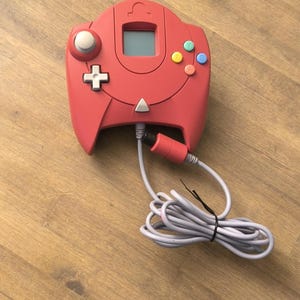 Peut inclure: Une manette rouge Nintendo 64 avec un cordon gris. La manette possède un petit écran, un joystick et plusieurs boutons.