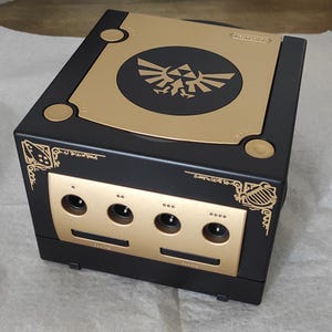 May include: Black and gold Nintendo GameCube console with a Zelda theme. The console has a gold top with a black circle and a Zelda crest. The front panel and decorative accents are also gold.