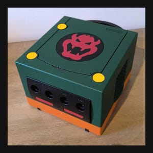 Op de afbeelding: Een op maat geschilderde Nintendo GameCube-console in groen en oranje, met een Bowser-embleem erop. De console heeft gele knoppen en een zwart voorpaneel met controllerpoorten. Een uniek gaming verzamelobject.