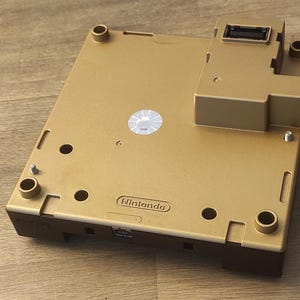 Puede incluir: Consola GameCube de color dorado con una base negra y una pegatina que dice "Nintendo".