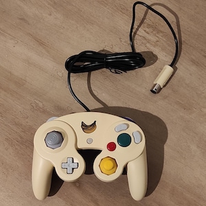 May include: Cream-coloured video game controller with a black cable and connector. The controller has a grey joystick, a directional pad, and coloured buttons in red, green, and yellow. A banana logo is visible.