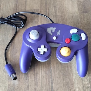 Op de afbeelding: Een paarse GameCube-controller met een witte voorplaat en een witte en grijze bedieningshendel. De controller heeft een gele C-stick, een groene Z-knop en een rode A-knop. De controller heeft een witte sticker aan de bovenkant met een silhouet van een geest.