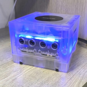 May include: A transparent blue GameCube console with a glowing blue internal light. The console has four controller ports and two memory card slots. The top of the console has a black disc tray.