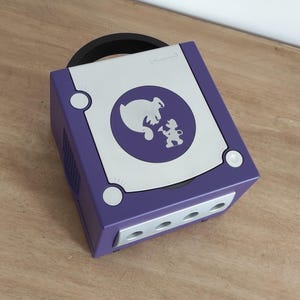 May include: A purple Nintendo GameCube console with a silver faceplate featuring a silhouette of a ghost and a character holding a paintbrush. The console has a handle on the top and a memory card slot on the front.
