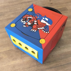 May include: A custom-painted Nintendo GameCube console. The top is split diagonally, with blue and red sections. The blue side features a red and white creature, while the red side has a blue and white creature. Yellow buttons and a yellow front panel complete the design.