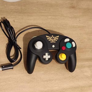 Może przedstawiać: Czarny kontroler GameCube z złotym symbolem Triforce na górze. Kontroler ma biały przycisk po lewej stronie, żółty przycisk po prawej stronie i niebieski przycisk na górze.