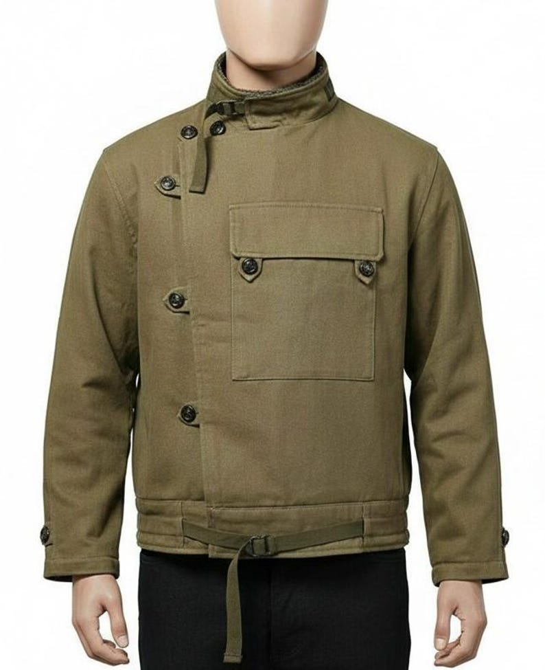 May include: Olive green jacket with a high collar, button closures, and a front pocket. The jacket features a belt at the waist and button details on the sleeves. The jacket is made of a durable fabric.