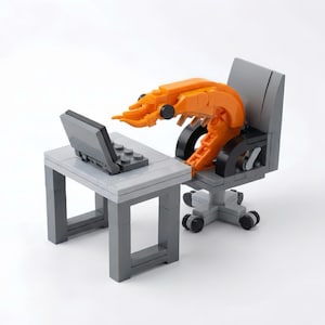 May include: A Lego model depicting an orange shrimp at a grey desk, using a grey laptop. The shrimp is in a grey office chair. The scene is set against a white background.