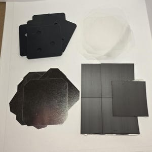 May include: An assortment of black and silver-colored square and octagonal shapes, along with clear, circular, thin sheets. The black squares have holes. The items are arranged on a white surface.
