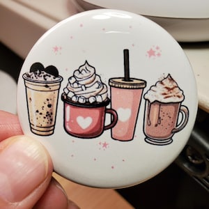 May include: A white button featuring four illustrated beverages: a coffee drink with chocolate, a hot chocolate with marshmallows, a pink drink with a straw, and a mocha. Pink stars and hearts decorate the button.