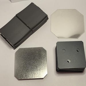 May include: An assortment of metal components, including stacks of square and octagonal pieces. The pieces are in shades of gray and silver, with some having circular cutouts. The surfaces show a brushed metal texture.