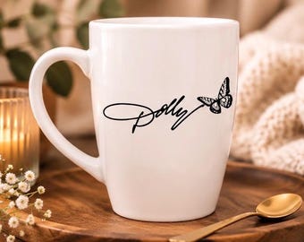 Puede incluir: Una taza de cerámica blanca con un diseño gráfico negro en la parte frontal. El diseño consiste en la palabra Dolly escrita en una fuente cursiva, seguida de una ilustración detallada de una mariposa con alas estampadas a la derecha.