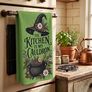 Op de afbeelding: Een felgroene wafelstructuur theedoek met een zwarte heksenhoed, een zwarte ketel en bloemaccenten. De tekst luidt "THE KITCHEN IS MY CAULDRON". Het ontwerp bevat diverse kruiden en witte madeliefjes rondom de centrale grafische elementen.