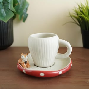 Könnte beinhalten: Eine weiße Kaffeetasse aus Keramik mit vertikalen Rippendetails, die auf einer Untertasse mit rotem Rand und weißen Tupfen steht. Eine kleine Eichhörnchenfigur sitzt auf der Untertasse. Die Tasse und Untertasse stehen auf einer Holzoberfläche.
