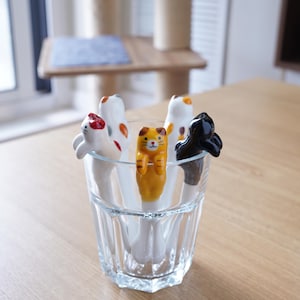 May include: A clear glass containing several ceramic spoons with cat designs. The spoons have white handles and feature orange, black, and white cat figures with red accents. The spoons are arranged in a clear, faceted glass.