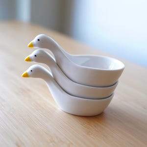 May include: Three white ceramic goose-shaped serving dishes stacked on top of each other. Each dish features a curved neck and a bowl. The goose heads have yellow beaks and small black eyes. The dishes are on a light-coloured wooden surface.