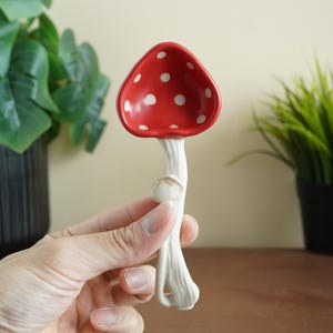 May include: A ceramic mushroom-shaped spoon rest. The spoon rest has a red cap with white polka dots and a white stem. The spoon rest is designed to hold spoons and other cooking utensils. The mushroom spoon rest is a decorative kitchen accessory.