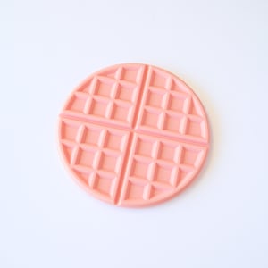 Puede incluir: Un posavasos de color rosa en forma de gofre. El posavasos está hecho de plástico liso y duro y tiene un patrón de gofre en relieve.