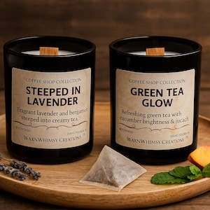 May include: Two black candles with wooden wicks and labels that read "Steeped in Lavender" and "Green Tea Glow." The candles sit on a wooden tray with lavender sprigs, a tea bag, a peach slice, and mint leaves. The candles are part of the "Coffee Shop Collection."