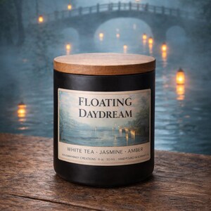 May include: A black candle jar with a wooden lid, labeled "Floating Daydream." The label features a serene landscape scene. The candle is on a wooden surface with a blurred background of a bridge and water.