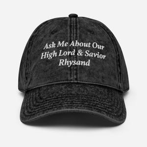 May include: A dark grey baseball cap with the text "Ask Me About Our High Lord & Savior Rhysand" embroidered in white. The cap has a curved peak and a button on top. The fabric has a faded, vintage look.