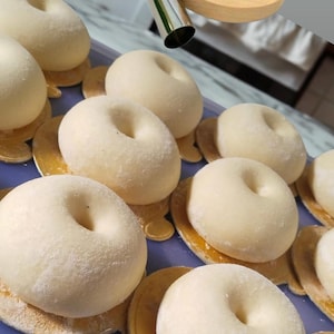 May include: An overhead shot shows a tray of raw, round donuts with a hole in the center, dusted with flour. A wooden dreidel and a metal pastry tube are in the background. The donuts sit on small, gold-colored, decorative bases.