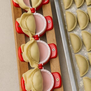 May include: A close-up shot of a wooden board with three red and white dumpling makers, each holding a filled dumpling. A metal tray with several uncooked dumplings is to the right. The dumplings are a light yellow color.