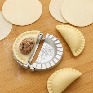 May include: A silver dumpling maker with a floral design is shown with a dumpling in progress, filled with ground meat. Several uncooked dumpling wrappers and finished dumplings are on a wooden surface.