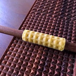 May include: A close-up of a yellow, textured food item, possibly a pastry, resting on a dark wooden stick. The stick is placed on a dark brown, patterned surface with a repeating geometric design. The food item has a waffle-like texture.