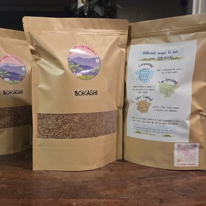 May include: Three brown paper bags of Bokashi, a composting product. Each bag has a clear window showing the contents. The bags have a circular label with a mountain scene and the text "Bokashi". One bag has a printed sheet with instructions.