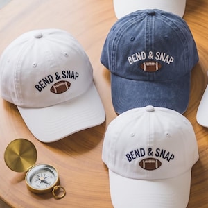 May include: Five baseball caps arranged in a circle, four white and one blue, each featuring the embroidered text BEND & SNAP above a brown football graphic. The caps have a structured crown, a curved brim, and a button on the top center.