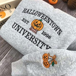 May include: A gray sweatshirt with the text "Halloweentown University" and a pumpkin graphic. The sleeve features a pumpkin and cherry design. The sweatshirt has the year "1998" printed on it.