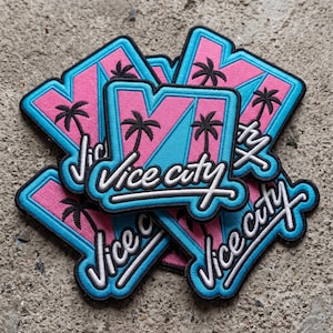 May include: A stack of embroidered patches featuring a pink and light blue geometric background with black palm tree silhouettes. The text Vice City is stitched in white cursive script across the center of each patch with a black border.